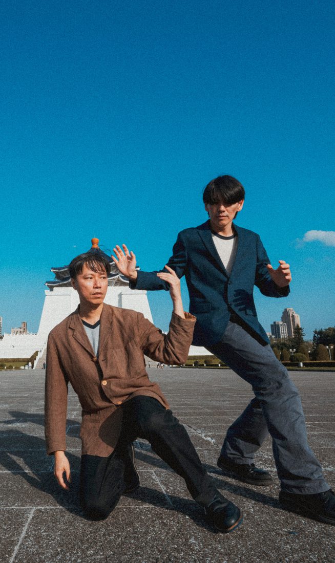 Two performers pose in a large public square under a clear blue sky. One kneels on the ground while the other stands beside them, both holding stylised dance positions. A large monument and city buildings are visible in the background.