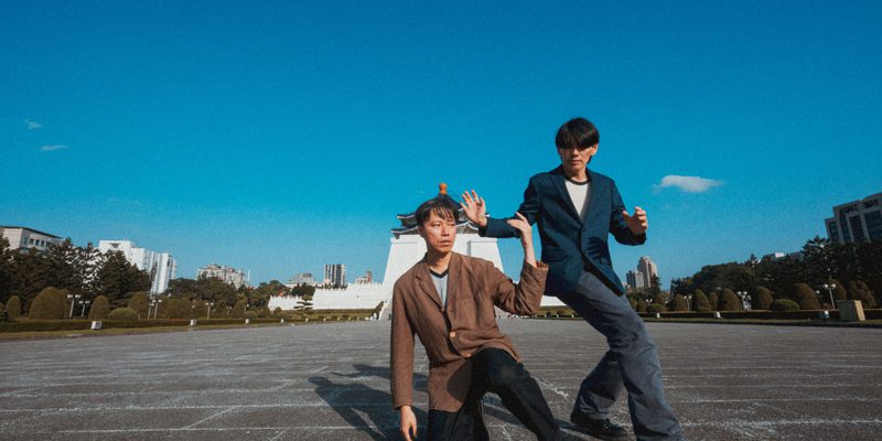 Two performers pose in a large public square under a clear blue sky. One kneels on the ground while the other stands beside them, both holding stylised dance positions. A large monument and city buildings are visible in the background.