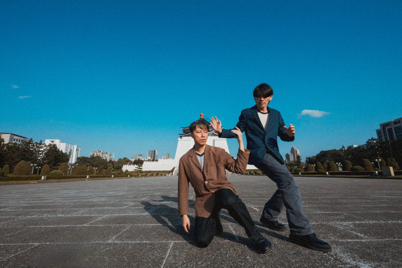 Two performers pose in a large public square under a clear blue sky. One kneels on the ground while the other stands beside them, both holding stylised dance positions. A large monument and city buildings are visible in the background.