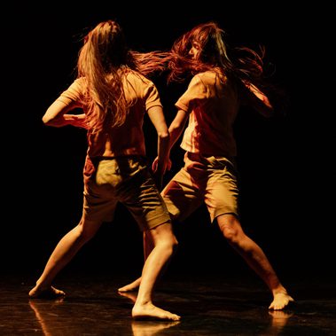 Two performers move in mirrored unison, their long dark hair flying around them. They are facing each other on a dark, glossy stage.