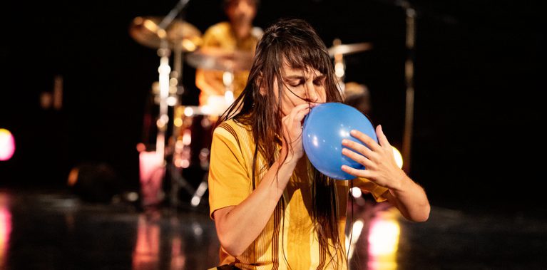 A dancer with long dark hair kneels and blows into a blue balloon, with a drummer visible in the blurred background.