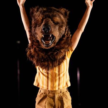 A performer stands with their arms raised, wearing a yellow striped shirt, similarly coloured trousers, and a full, furry brown bear head mask with an open mouth and bared teeth.
