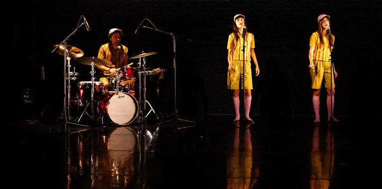 A drummer sits at a red drum kit on the left side of a dark, reflective stage. On the right, two performers stand with microphones, all three in matching outfits, yellow tops and bottoms as well as pink knee-high socks.