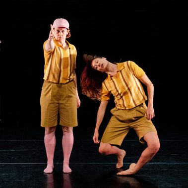 One performer stands confidently and points directly forward, while the other performer next to them is in a deep, contorted pose, with their head tilted far back and their body leaning to the side.