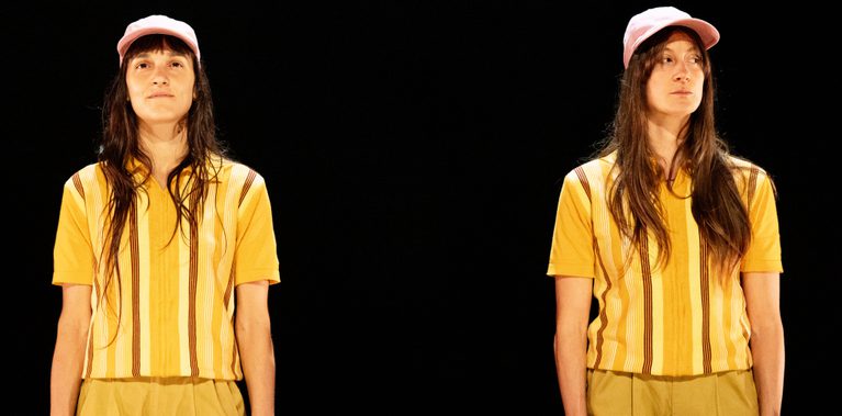 Two almost identical dancers with long dark hair wearing matching yellow striped shirts, tan shorts, and pink hats stand side-by-side, the performer on the left has a soft almost smile while the one on a right a more mischievous look. They stand together against a black background.