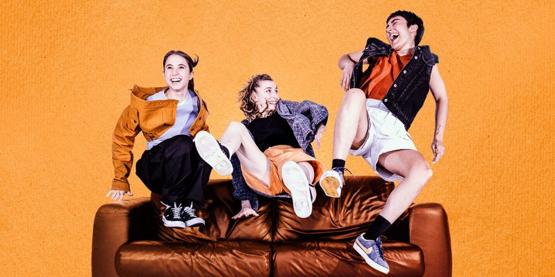 Three dancers leap up onto a brown leather sofa, smiling big warm smiles at each other, clearly they are close friends. Their outfits are all 90s inspired in shades of orange and red. The background of the image is an orange, textured like paper.