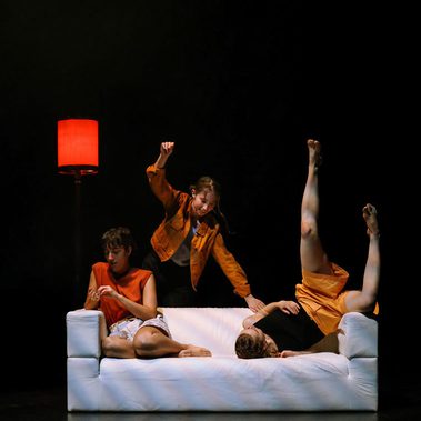 Three dancers tumble together on a plain white sofa. On the far left, the dancer lies with her torso horizontal on the sofa but with her legs suspended up in the air. The dancer in the centre smiles down at her as she leaps over the back of the sofa. On the left, a third and final dancer is seated with her feet tucked up under her. She is deep in thought, removed from the other two, staring into her hands. The soft red glow of a lamp shade is visible behind them on an otherwise black stage.
