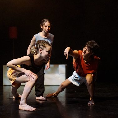 A dancer lunges towards the camera and points to a spot on a dark dance floor. On her left, another dancer mirrors her lunch and looks inquisitively at her pointing finger. Behind them both, a third dancer tries to see what they're looking at.