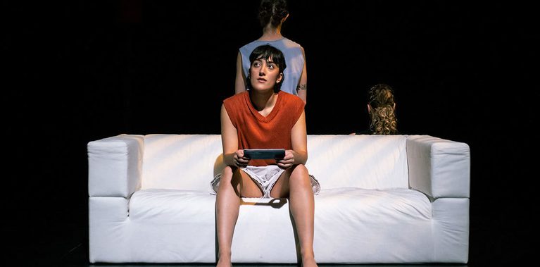 A dancer with short, dark hair and a rust coloured knitted vest sits on a plain white sofa. She holds a plane ticket in her hands and looks thoughtfully off into the distance, as if considering what meaning the ticket has on her current life. Her two friends are perched behind the sofa with their backs to her, not seeing the ticket.