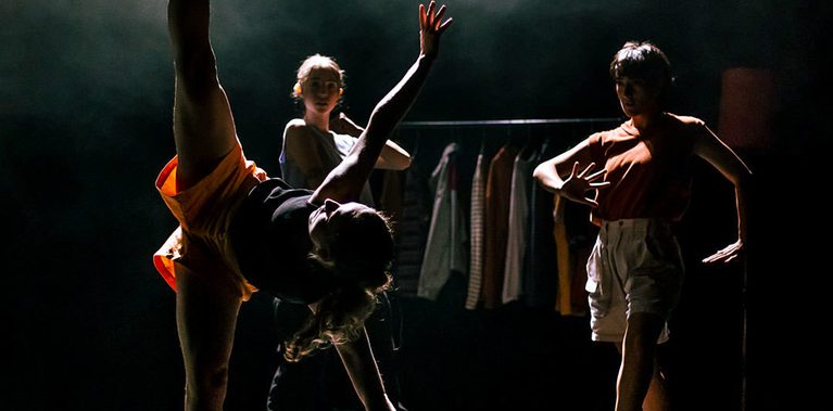 A dancer stands with one leg on a dark stage, her other in a straight line above her in the splits. Her torso is parallel to the floor, her arms outstretched. Behind her are two other dancers, back lit by a high contrast light, shining through haze.