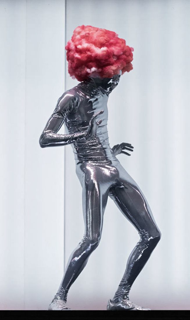 A shiny, silver being wearing a pink candy floss wig dances against the white, lit interior of the shipping container.