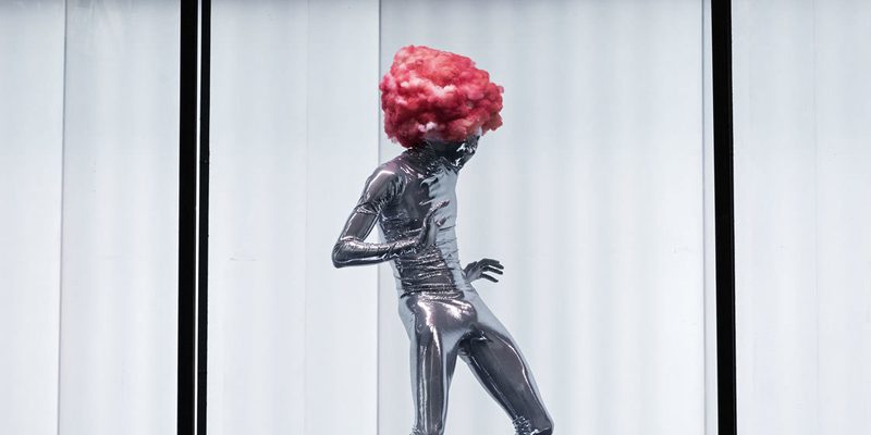 A shiny, silver being wearing a pink candy floss wig dances against the white, lit interior of the shipping container.