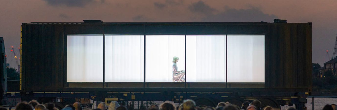Set against the sky at dusk, a captive audience all wearing headphones are seated in front of a large, dark shipping container, mounted on the bed of a 40ft truck. The long side of the container has been removed revealing a single silver being, crouched on the floor.