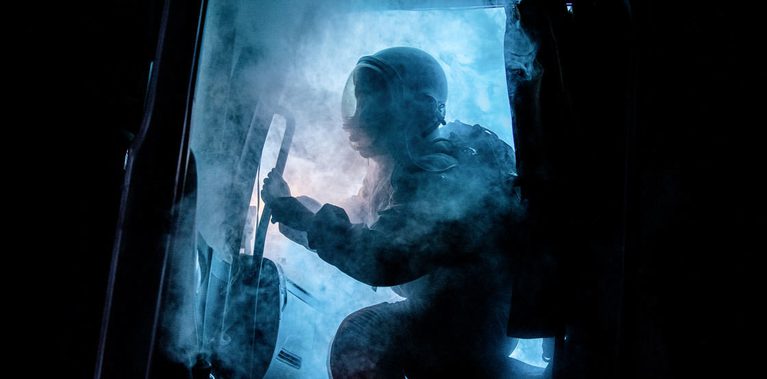 The cabin door of a truck opens, spewing eerie blue smoke. Silhouetted by a bright blue light that cuts through the darkness of night, a figure wearing an astronaut uniform begins to emerge.