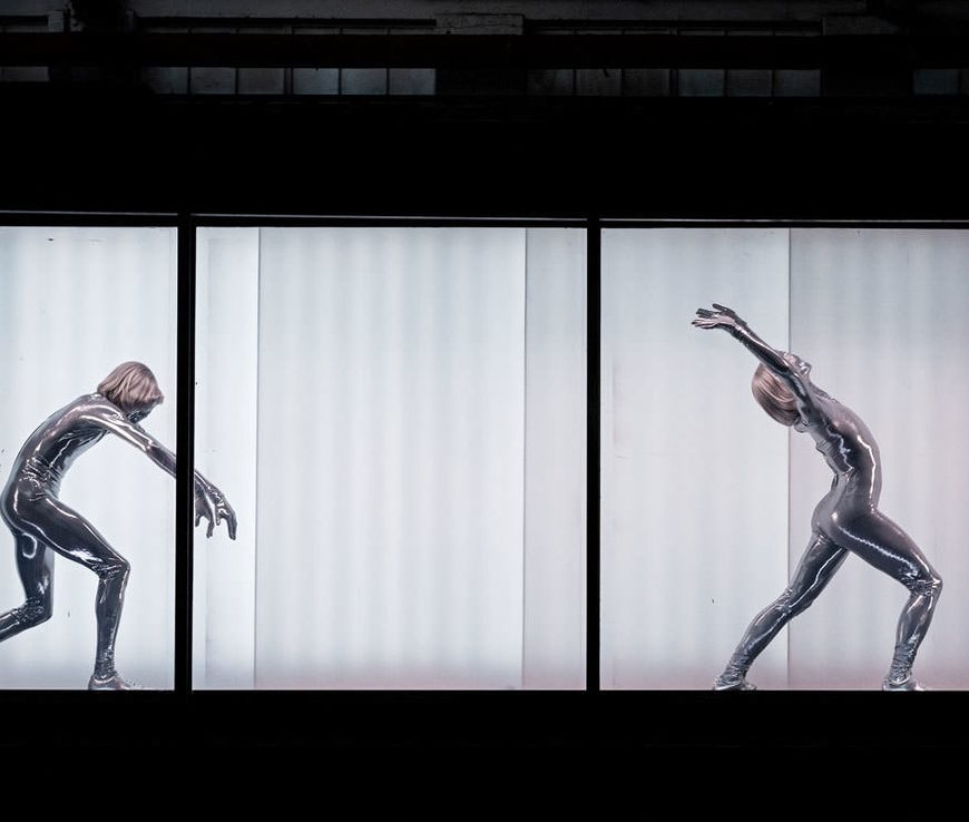 Two shiny, silver beings dance elegantly in uniform. Both facing to the right as the move along the hidden travelator inside the shipping container, the being on the left bends down, in an opposite motion to the being on the right who extends their arms above their head and arches back.