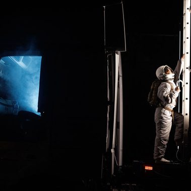 Against the dark of night, an astronaut begins their ascent on a ladder, mounted on the side of the shipping container, in the gap between the cabin of the truck and the container it is attached to. The cabin is lit up, spewing blue smoke from the open door.