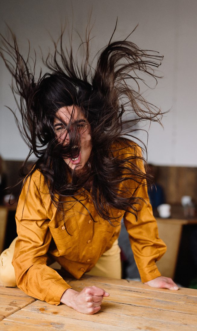 A woman wearing an orange shirt bangs her fist angrily on the table.