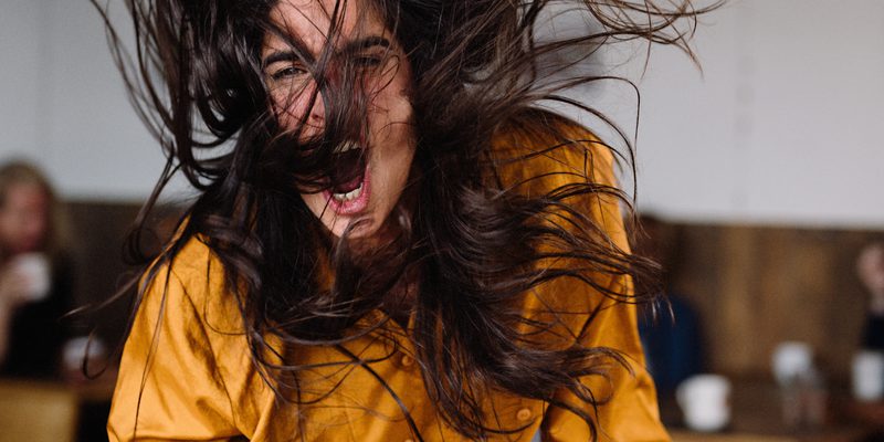A woman wearing an orange shirt bangs her fist angrily on the table.