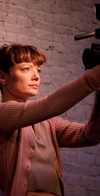 A female performer is standing if front of a white bricked wall holding a camera which she has turned in her direction facing her and lighting her face.