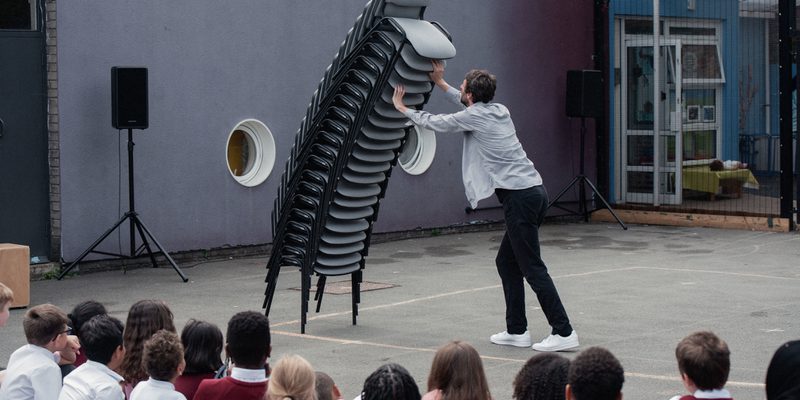 A man in a light denim shirt, dark jeans and white trainers holds a leaning tower of stacked office chairs as they lean towards him. They are stacked up higher than his head, and although they look like they might come crashing down to earth at any second, they seem suspended in the air. In the foreground is an audience of primary school students completely entranced.