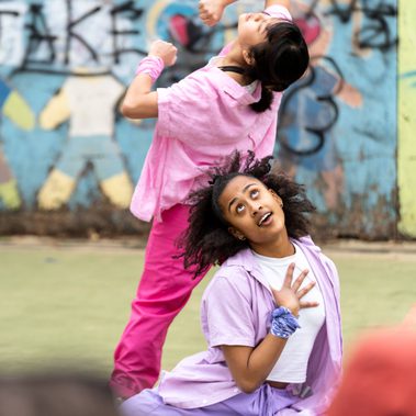 Lisa, a Southeast Asian, female dancer of small build, with black shoulder-length hair and black eyes, wearing a white shirt, dark pink trousers and a light pink button-up shirt, with a pink bandana around her wrist, dances with her back to the audience, her bending backwards with her fists at her chest and her elbows held high. Kassi, who has brown skin and afro-curly hair, wearing light purple trousers, a light purple shirt with a white vest underneath, is seated behind Lisa. Her hand is held to her chest, fingers splayed, her eyes looking up to the sky. The audience are visible in the foreground.