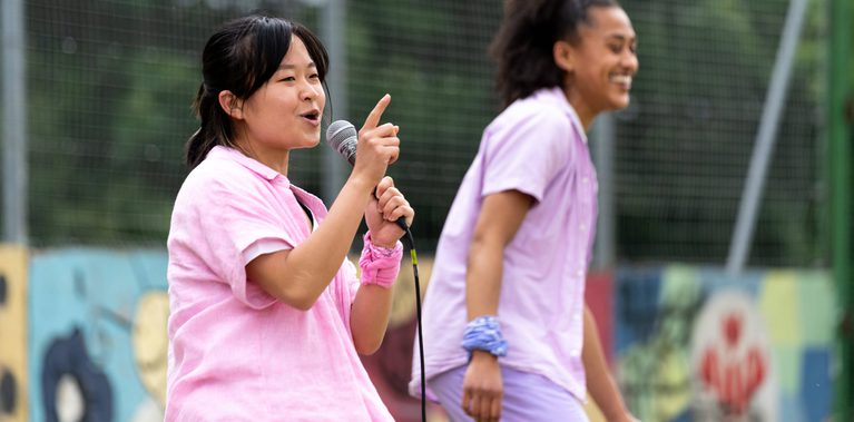 Lisa a Southeast Asian, female dancer of small build, with black shoulder-length hair and black eyes, wearing a white shirt, dark pink trousers and a light pink button-up shirt, with a pink bandana around her wrist, talks into a microphone and gestures with her figure. Kassi, who has brown skin and afro-curly hair, wearing light purple trousers, a light purple shirt with a white vest underneath, stands in background
