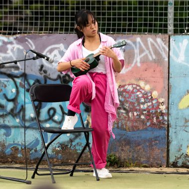 Lisa, a Southeast Asian, female dancer of small build, with black shoulder-length hair and black eyes, wearing a white shirt, dark pink trousers and a light pink button-up shirt, with a pink bandana around her wrist, plays a small green ukulele close to a microphone, one knee raised on a chair.