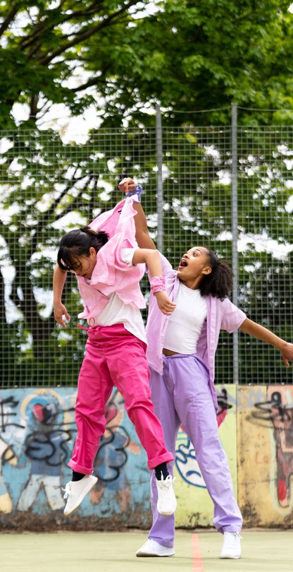 Lisa, a Southeast Asian, female dancer of small build, with black shoulder-length hair and black eyes, is being thrown in the air by the scruff of her neck by Kassi who is much taller! Lisa is wearing a white shirt, dark pink trousers and a light pink button-up shirt, with a pink bandana around her wrist. Kassi, who has brown skin and afro-curly, is wearing light purple trousers, a light purple shirt with a white vest underneath. She is wearing white trainers.