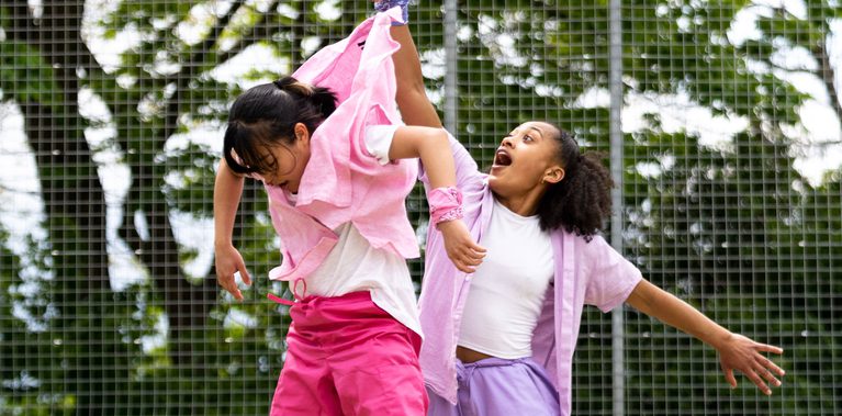 Lisa, a Southeast Asian, female dancer of small build, with black shoulder-length hair and black eyes, is being thrown in the air by the scruff of her neck by Kassi who is much taller! Lisa is wearing a white shirt, dark pink trousers and a light pink button-up shirt, with a pink bandana around her wrist. Kassi, who has brown skin and afro-curly, is wearing light purple trousers, a light purple shirt with a white vest underneath. She is wearing white trainers.