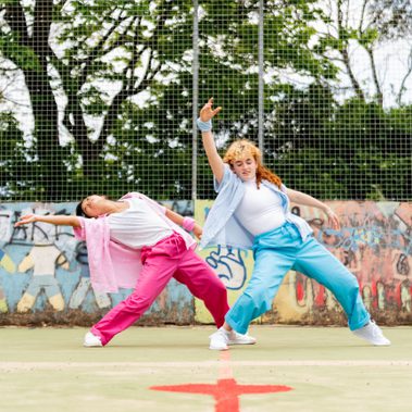 Lisa, a Southeast Asian, female dancer of small build, with black shoulder-length hair and black eyes, dances with her legs apart and her left arm and torso parallel to the ground. She is wearing a white shirt, dark pink trousers and a light pink button-up shirt, with a pink bandana around her wrist. Rose copies the shape of Lisa’s dance but has her hand pointed upwards. She has pale freckled skin, blue eyes, ginger curly hair, is small in height and wearing bright blue trousers with a pale blue shirt.