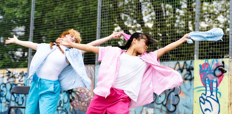 Lisa, a Southeast Asian, female dancer of small build, with black shoulder-length hair and black eyes, wearing a white shirt, dark pink trousers and a light pink button-up shirt, dances with her arms wide, looking towards the blue bandana in her right hand. Rose, who has pale freckled skin, blue eyes, ginger curly hair, is small in height and wearing bright blue trousers with a pale blue shirt, copies her in the background but with a pink bandana.