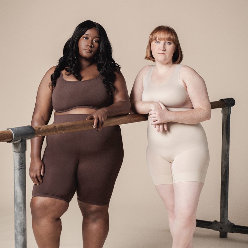 Two plus sized dancers wearing varying skin toned, body hugging shorts and bras, stand at a ballet bar. They stare out towards us, asking us to question what we see.