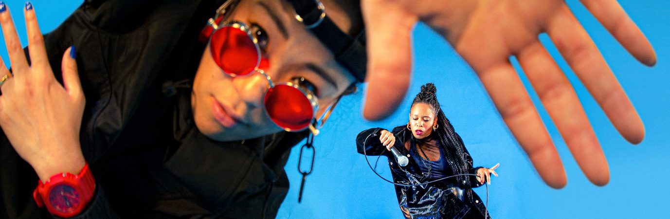 A dancer in all black trackies, a backwards black cap and red sunglasses and watch leads into the camera, their hand is up to the camera and is warped by a fish eye lense. In the background, another dancer in all black dances with a microphone in their hand. They slot into the space between the thumb and forefinger of the dancer in the foreground. The background is a bright electric blue.