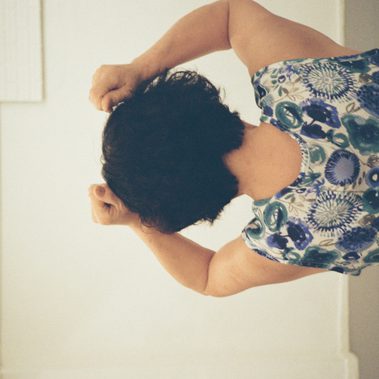 An elderly woman with short, dark and curly hair is seen from behind, with both hands raised near their head while wearing a floral-patterned clothing