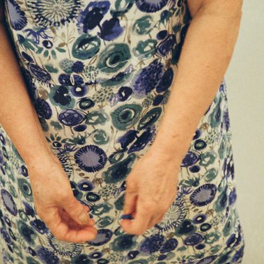 A close-up of an elderly woman’s arms and hands wearing a blue floral patterned dress, with a focus on the intricate flower designs and the position of the hands in subtle motion.