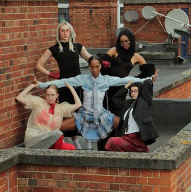 5 female dancers create a strong pose with a piercing gaze towards the camera. They wear a mixture of different textured and coloured clothing and are surrounded by brick structures.