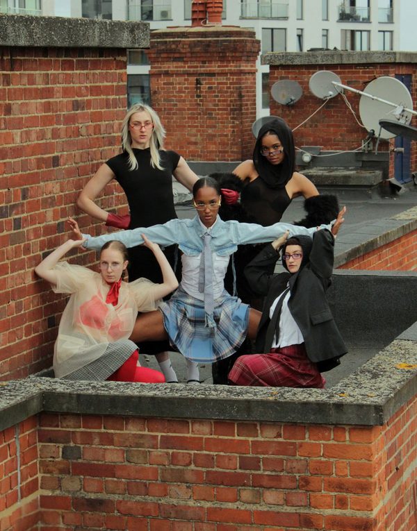 5 female dancers create a strong pose with a piercing gaze towards the camera. They wear a mixture of different textured and coloured clothing and are surrounded by brick structures.