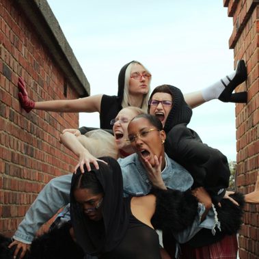 5 females are intertwined with animalistic facial expressions. All wear a mixture of textured and coloured clothing and stand between two brick structures.