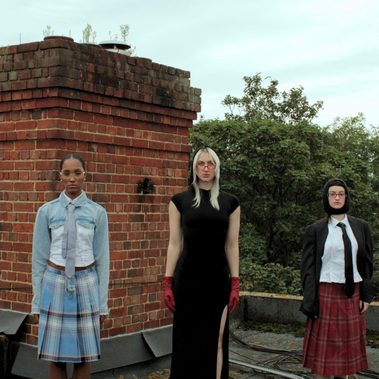 5 females stand in a line, looking strongly at the camera with a dead-pan expression and pose. All wear a mixture of textured and coloured clothing.
