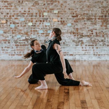 One dancer in front kneels on the floor and another dancer behind sits on their knee leaning to the side.