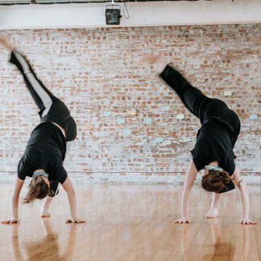 Two dancers side by side stand inverted with one leg toward the ceiling slightly out to the side.