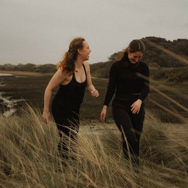 Two women, one with light brown hair and the other with dark brown hair, stand side by side laughing.