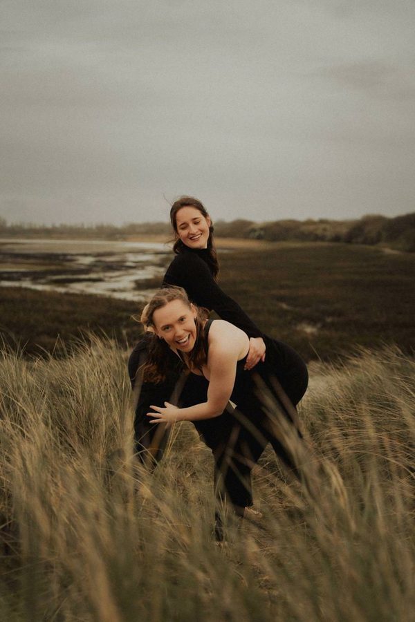 Two dancers with long hair, one leaning forward in the foreground, hold on to each other while smiling outside.