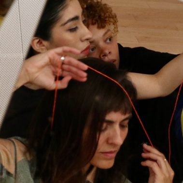 A white background with a square mirror reflection, cut off diagonally down across the right side. The reflection holds three faces of woman, layered and collaged with two in the background and one in the foreground. They look pensive, alert and serene with one woman holding a piece of red thread across her head from one hand to the other.