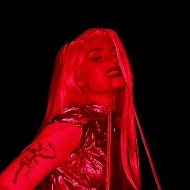 A young white woman with long blond hair wearing a silver bodice, wrapped in rope, smiles menacingly at the camera while biting a piece of rope. The image has a red hue with a black background.