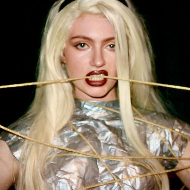 A young white woman with long blond hair wearing a silver bodice, wrapped in rope, stares into the camera while biting a piece of rope.