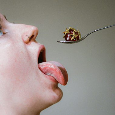 A glacé cherry covered with gold star pins is held out on a silver teaspoon. Below, a white person greedily sticks out their tongue, ready to devour.