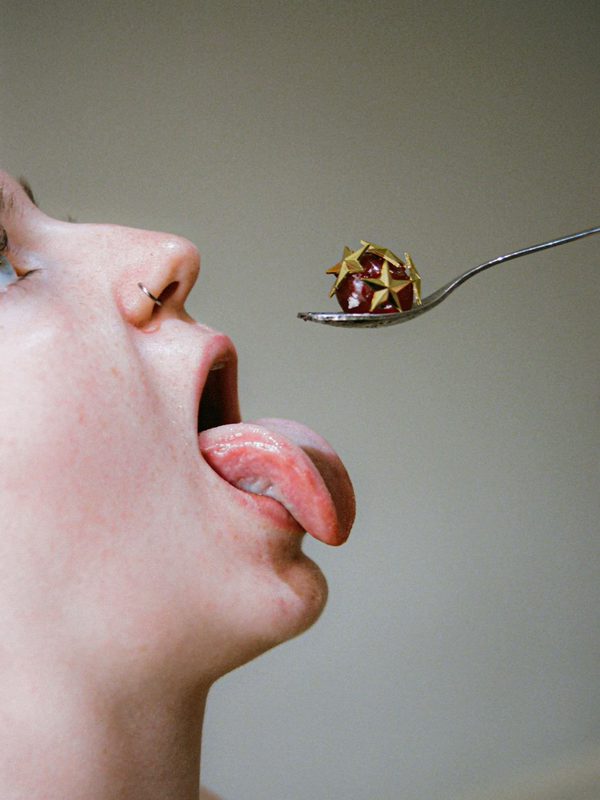 A glacé cherry covered with gold star pins is held out on a silver teaspoon. Below, a white person greedily sticks out their tongue, ready to devour.