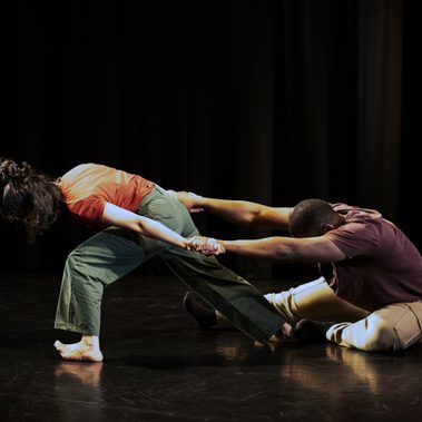 A performer sits on the floor with head bent, and is being dragged by the other performer, with her hands stretched backwards, and making a big effort to drag.