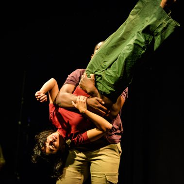 A performer is being swung around by the other with his arms around her, she is mid-turn and her body is a diagonal line in the air.